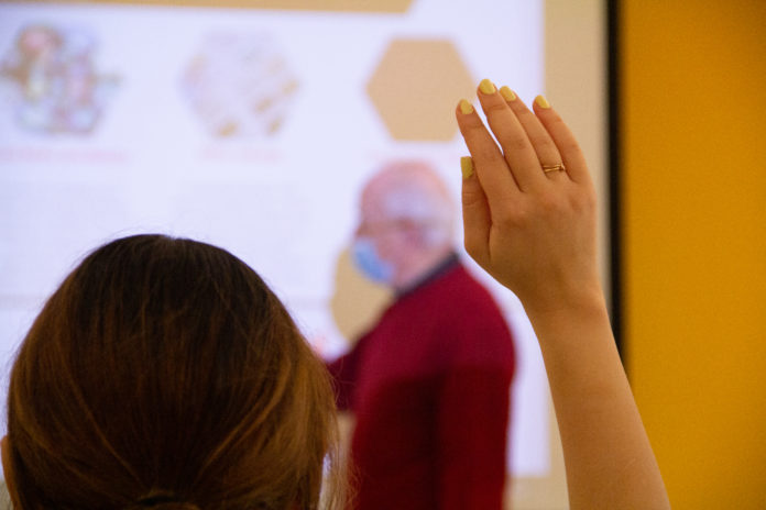 the photo is of the back of a student's head, the back of their raised hand in the foreground. The teacher points at a board, blurred out in the background.